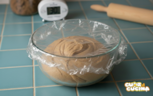 Panetto di pasta frolla alla cannella e miele in ciotola coperta da pellicola, pronto per il riposo in frigo.