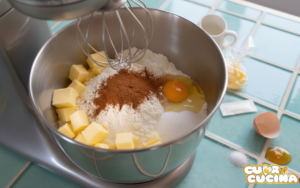 Ingredienti per pasta frolla alla cannella nella ciotola della planetaria: farina, burro a cubetti, uovo e spezie per i biscotti Musicarelli.